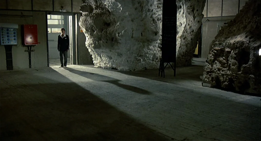 Intervista (1987) movie still: wide — A wide industrial space features large, unrefined rock formations and concrete flooring. A lone man stands in…
