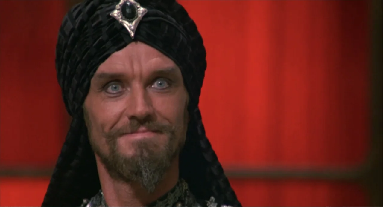 Hercules (1983) movie still: closeup — A figure stands in a room with a bright red wall behind him. The man wears an elaborate turban and stares…