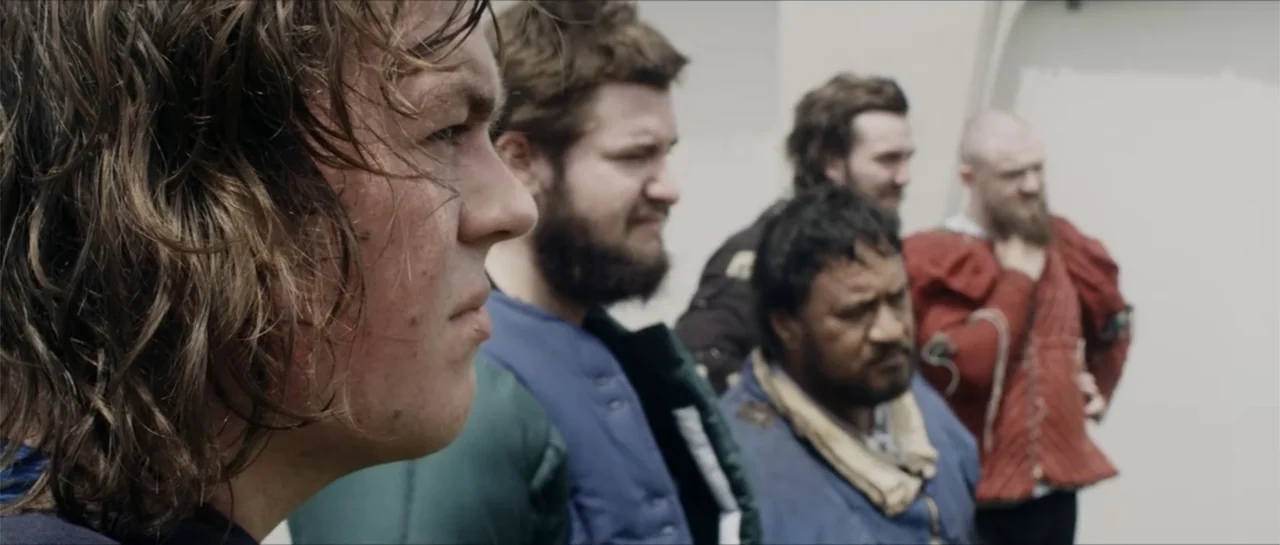 Bludgeon (2018) movie still: medium — A group of men stand in a plain indoor hall with light colored walls. Four men in medieval attire stand…