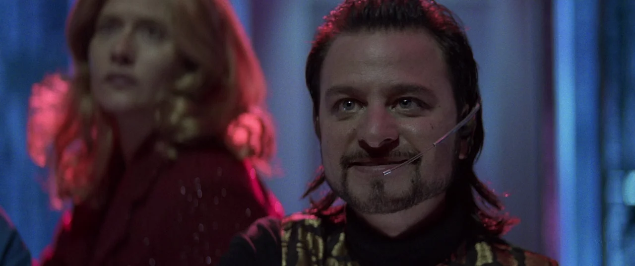 Hackers (1995) movie still: medium — A dimly lit room bathed in blue and magenta neon lighting effects. A man with a headset smiles while a blonde…