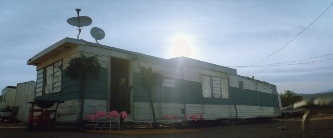 Gender Under Attack (2018) movie still: wide — An aging mobile home sits on a dusty lot under a clear midday sky. The structure is decorated with…