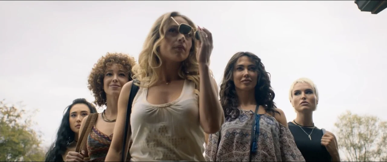 Risqué (2025) movie still: medium — Five women walk toward the camera in an outdoor setting under an overcast sky. The group consists of young women…
