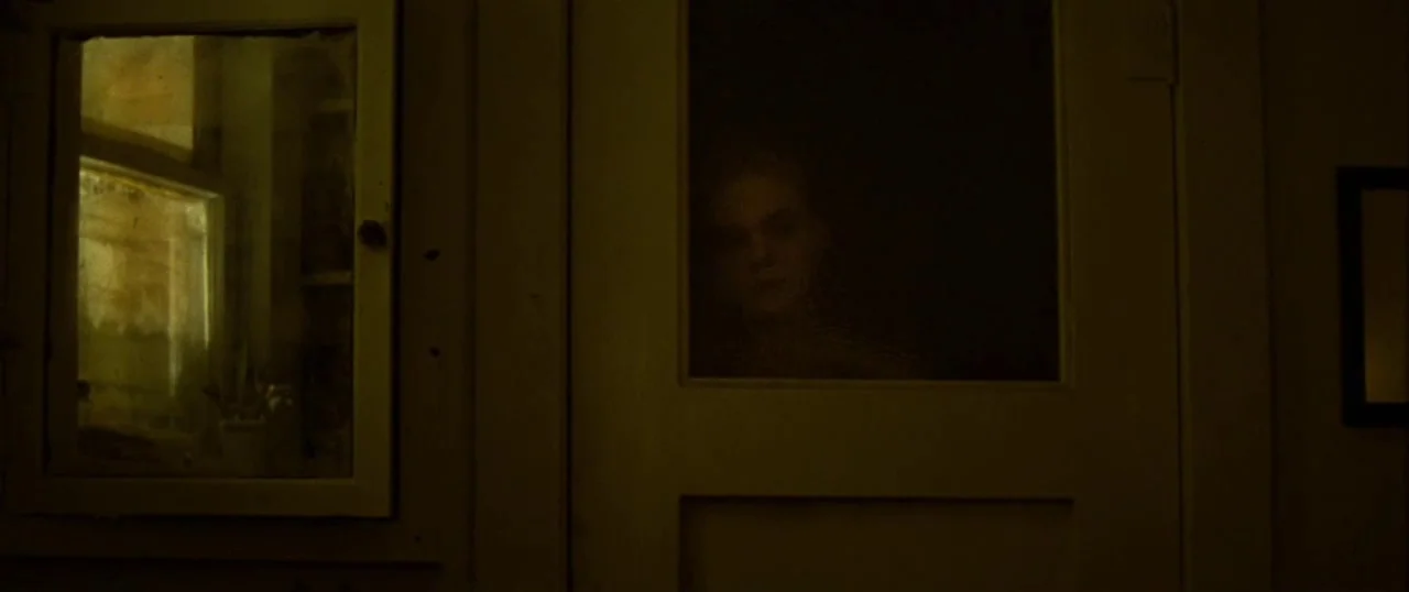 Low Down (2014) movie still: medium — A dimly lit room contains a white wooden door with a central glass pane. A young woman stands behind the glass…