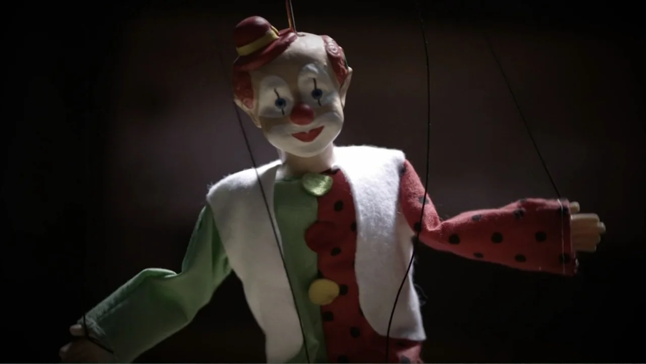 What Happened at Fells Acres? (2023) movie still: medium — A wooden clown marionette hangs suspended in a dark, empty interior space. The doll is…
