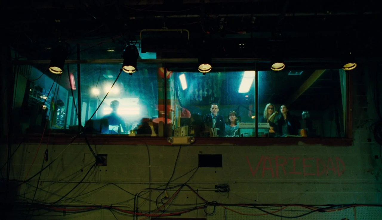 Our Brand is Crisis (2015) movie still: wide — An industrial control room features a large glass window overlooking a dimly lit workspace. Four…