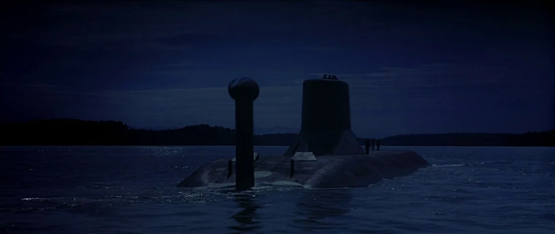The Hunt for Red October (1990) movie still: wide — A dark, partially submerged submarine moves through calm water under a deep blue sky. A dense…