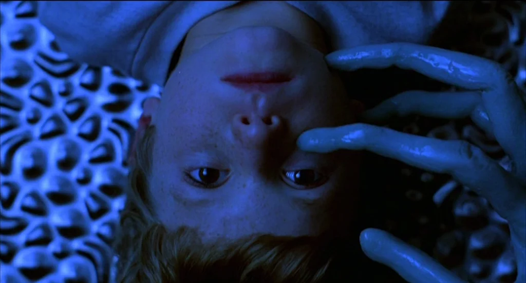 Mysterious Skin (2004) movie still: closeup — A boy lies on a textured metallic surface that reflects blue light. An elongated, smooth blue alien…