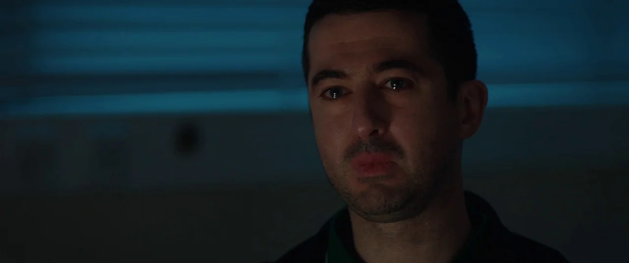 Journeyman (2017) movie still: closeup — A man stands inside a dimly lit space with blue light filtering through blinds. The man gazes toward the…