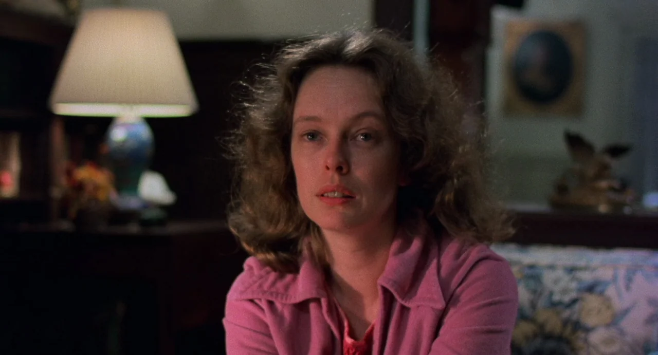 God Told Me To (1976) movie still: closeup — A dimly lit domestic interior features a woman sitting in the foreground. The background shows a table…