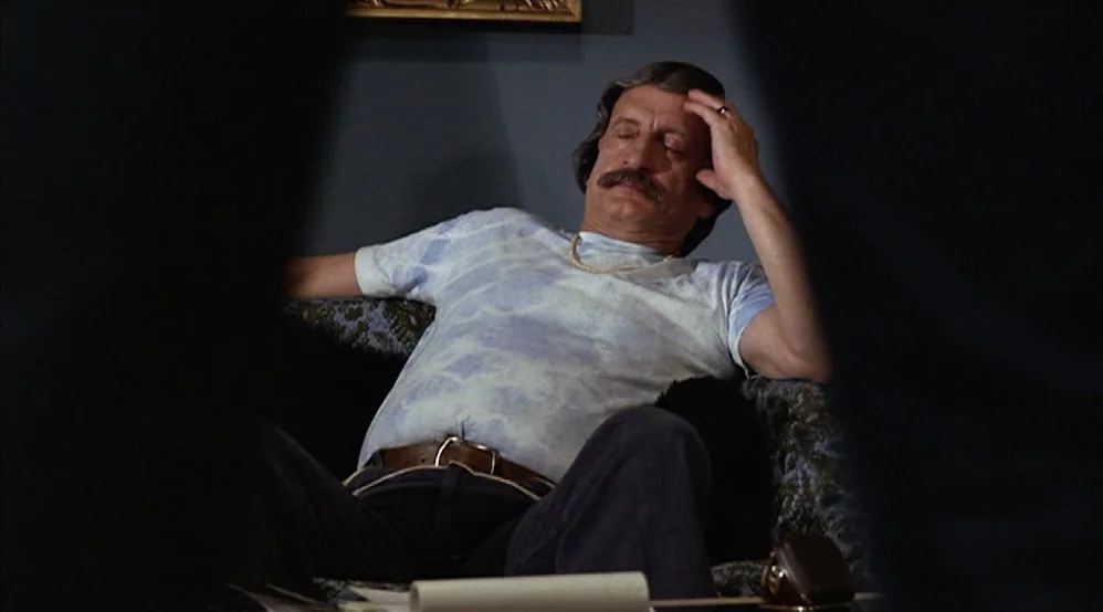 Hardcore (1979) movie still: medium — A man sits on a dark patterned couch against a solid grey wall. He rests his head in his hand while sitting in…