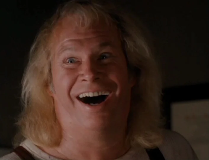 Head of the Family (1996) movie still: closeup — A man with shoulder-length blonde hair and a white shirt occupies a dark space. He smiles directly…