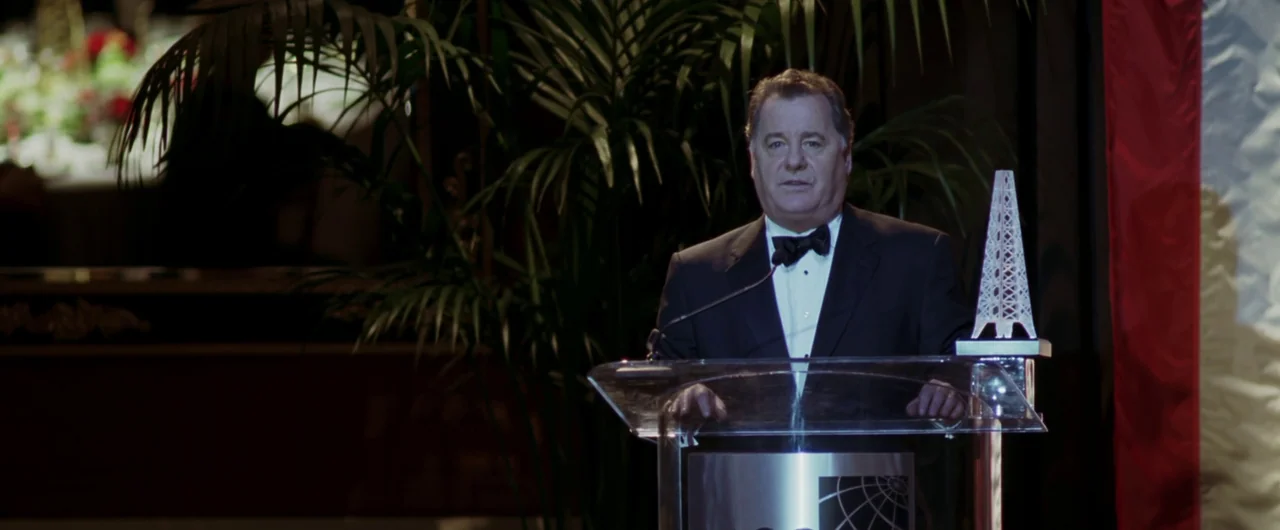 Syriana (2005) movie still: medium — An award ceremony held in a dark room with a large palm plant backdrop. A man in a tuxedo stands behind a…