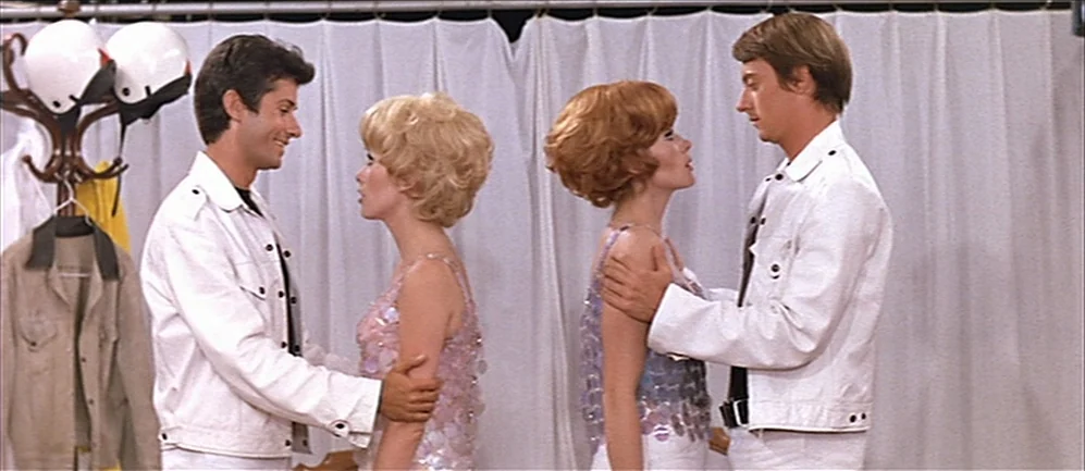 Les Demoiselles de Rochefort (1967) movie still: medium — A room with white curtains backdrop and a coat rack with helmets. Two couples stand facing…