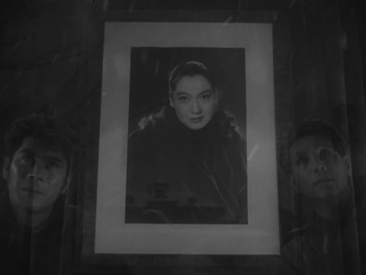The Idiot (1951) movie still: medium — A framed photograph of a woman rests against a wall, flanked by two men. The men gaze at the portrait in a…