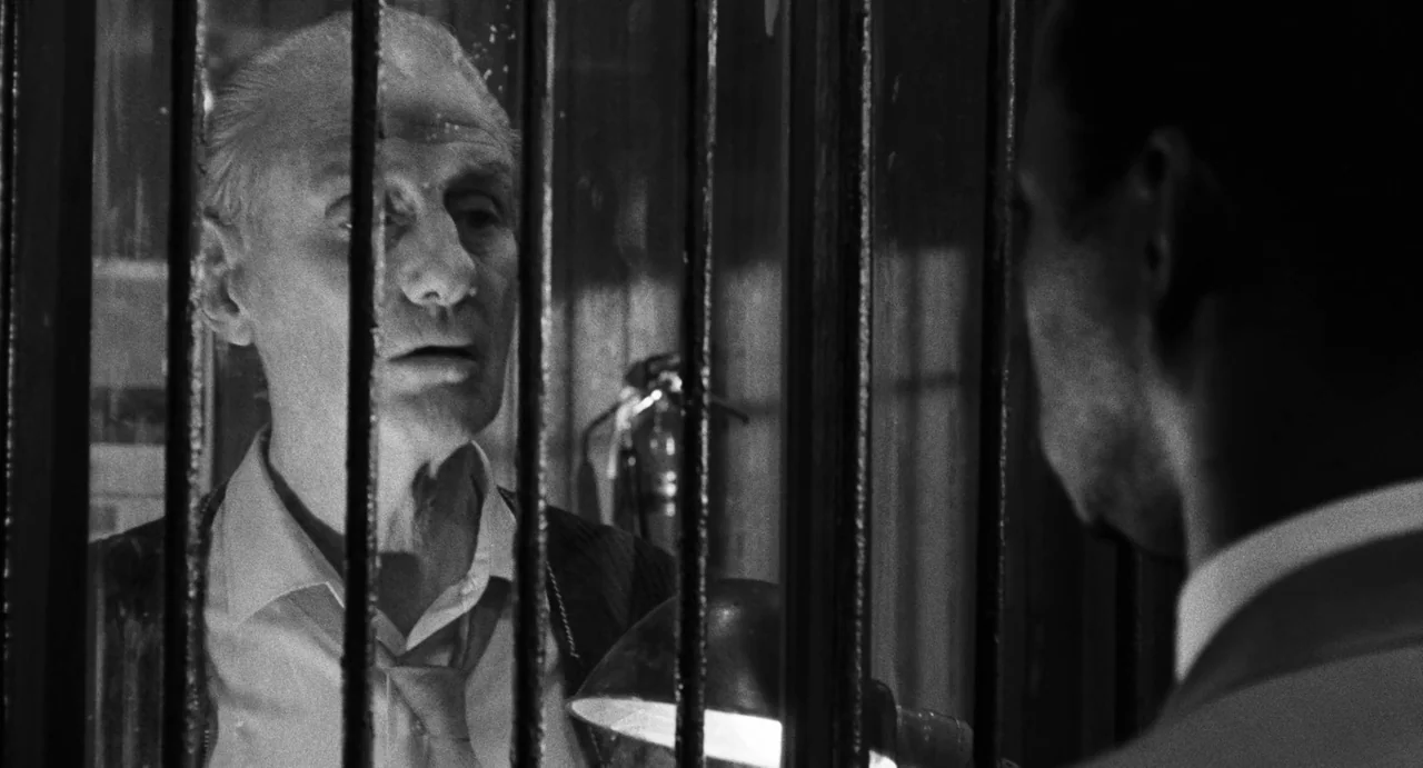 The Incident (1967) movie still: medium — A dimly lit room separates two men with heavy black metal bars. The older man sits behind the barrier…