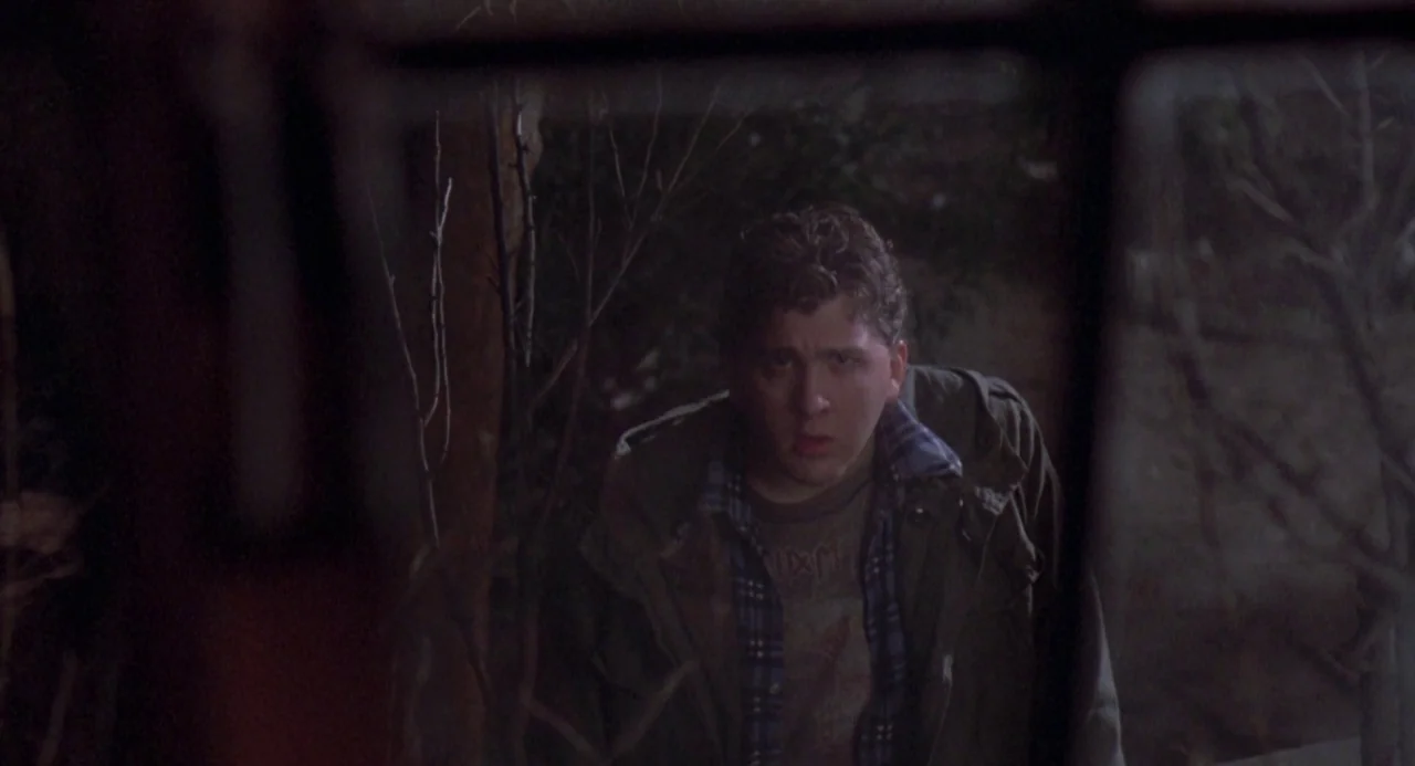 River’s Edge (1986) movie still: medium — A man stands in a dark, wooded area at night surrounded by bare branches. He looks toward the camera while…