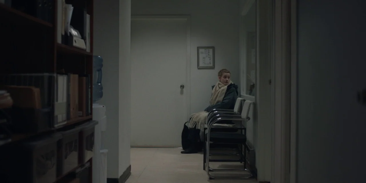 The Assistant (2019) movie still: wide — A dimly lit narrow hallway with a wood bookshelf and stack of chairs. A young woman sits alone on a metal…