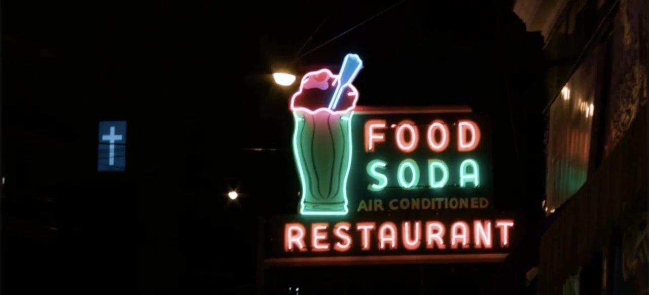 Hellbreeder (2004) movie still: wide — A dark urban exterior features a glowing neon sign for a restaurant at night. A small square light with a…