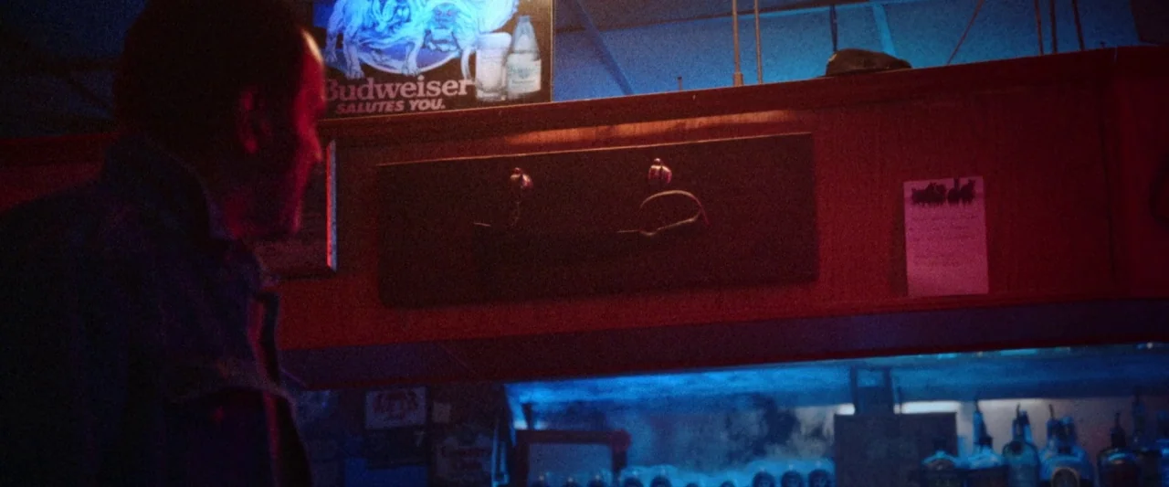 VFW movie still: medium — A dimly lit tavern with a wooden bar, red walls, and blue neon illumination. A man stands in the foreground, his profile…