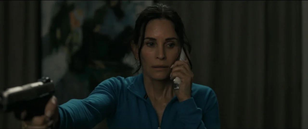 Scream VI (2023) movie still: medium — A dark living room interior with draped curtains visible behind a woman. A woman holds a firearm in one hand…