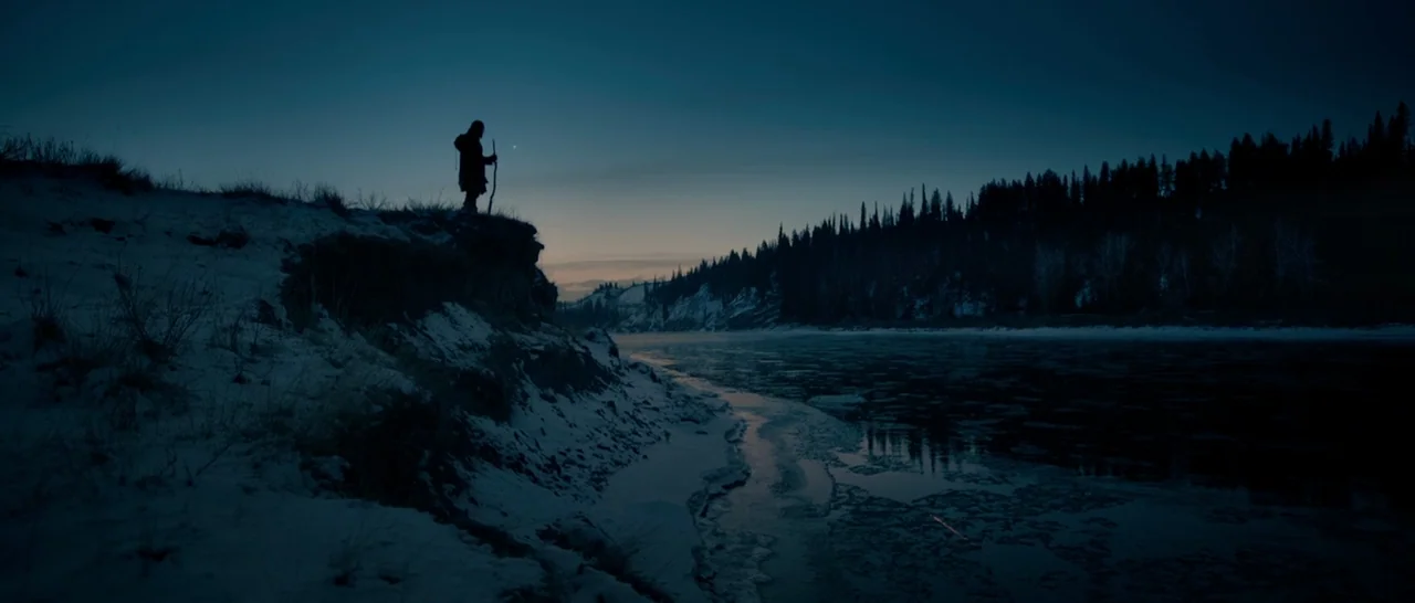 The Revenant (2015) movie still: wide — A snowy, darkened riverbank overlooks a cold, partially frozen river at twilight. A solitary hooded figure…