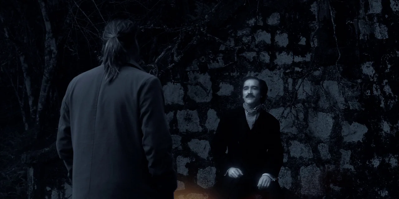 Twixt (2011) movie still: medium — A man stands before another man seated against an old, uneven stone masonry wall. The environment is dark and…