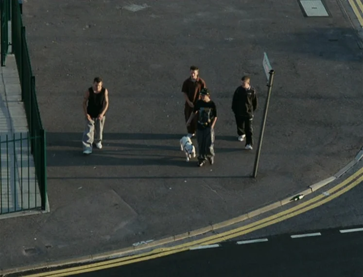 Fish Tank (2009) movie still: wide — A paved corner of an urban street with painted road markings and a signpost. Four young men walk across the…