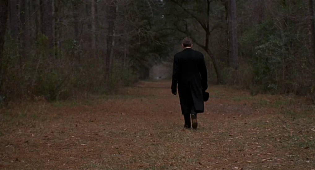 Miller’s Crossing (1990) movie still: fullbody — A quiet, narrow dirt trail cuts through a dense forest of leafless trees. A lone man in a dark coat…