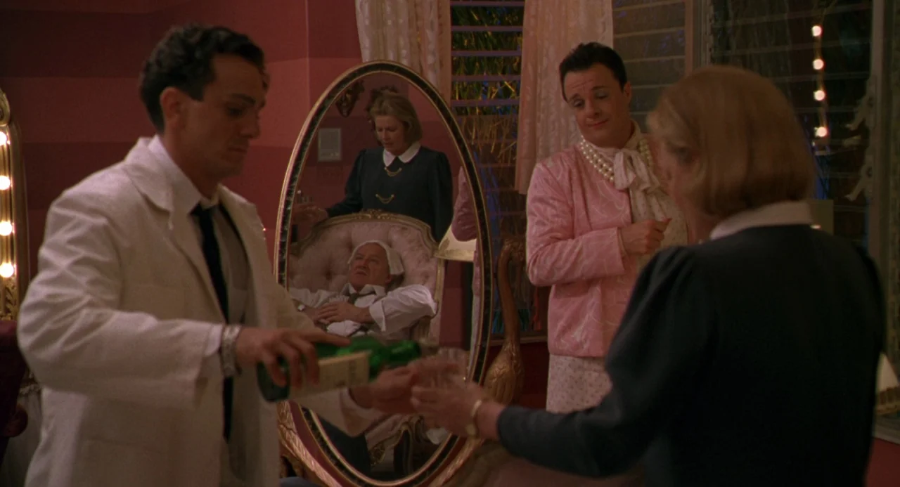 The Birdcage (1996) movie still: medium — A lavish dressing area with striped wallpaper and an ornate oval floor mirror. A man pours liquid into a…
