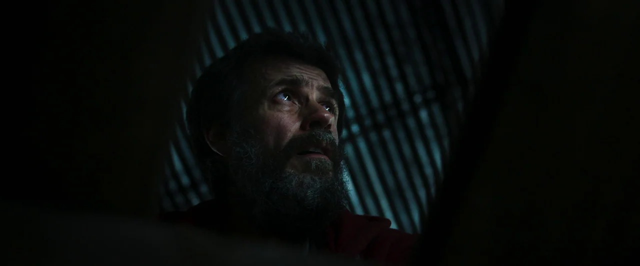 When Evil Lurks (2023) movie still: closeup — A man in a dark hoodie sits in a dimly lit, confined space. He gazes upward with a wide-eyed…