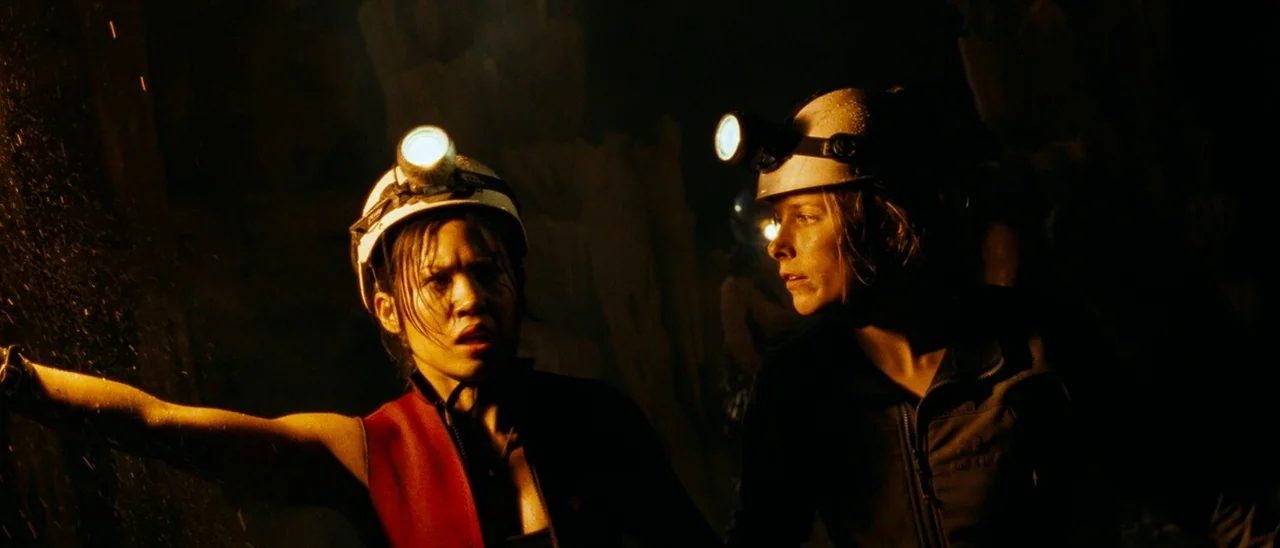 The Descent (2005) movie still: medium — Two women explore the dark, damp interior of a rugged cave system. They scan the shadows with headlamps,…
