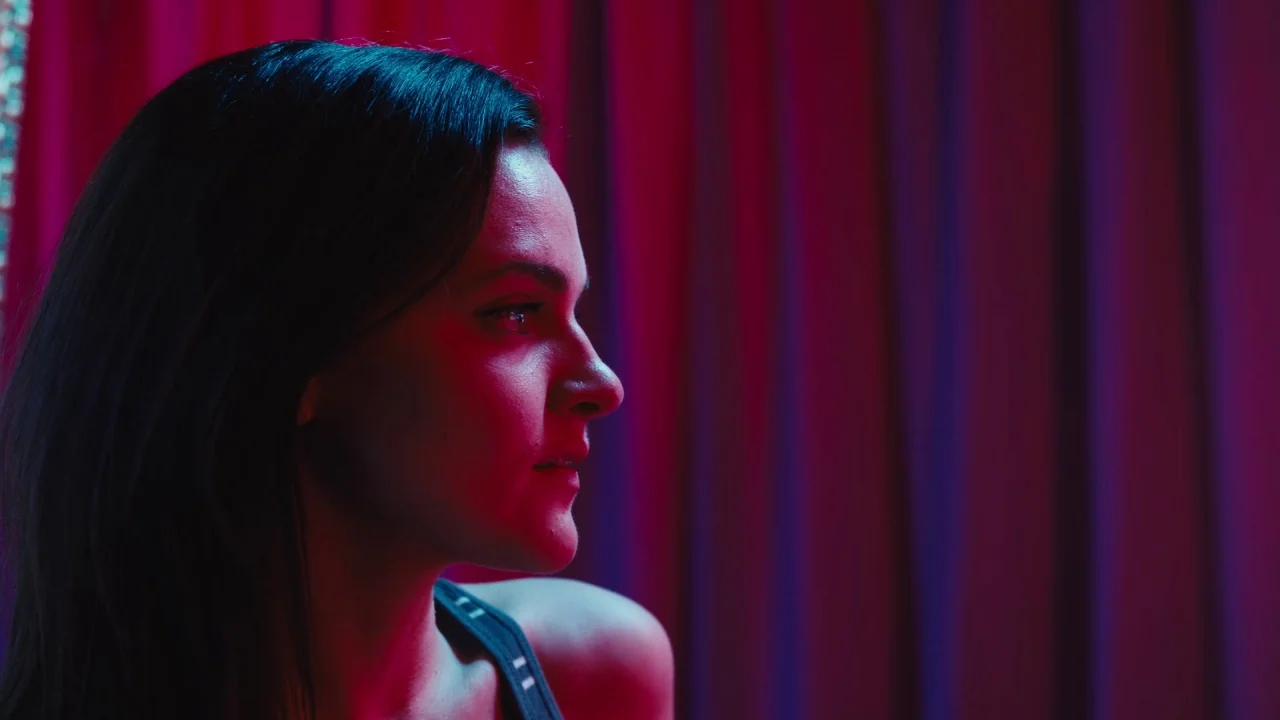 Cam (2018) movie still: closeup — A woman is positioned in front of heavy dark purple curtains in a room. She looks off-screen while illuminated by…