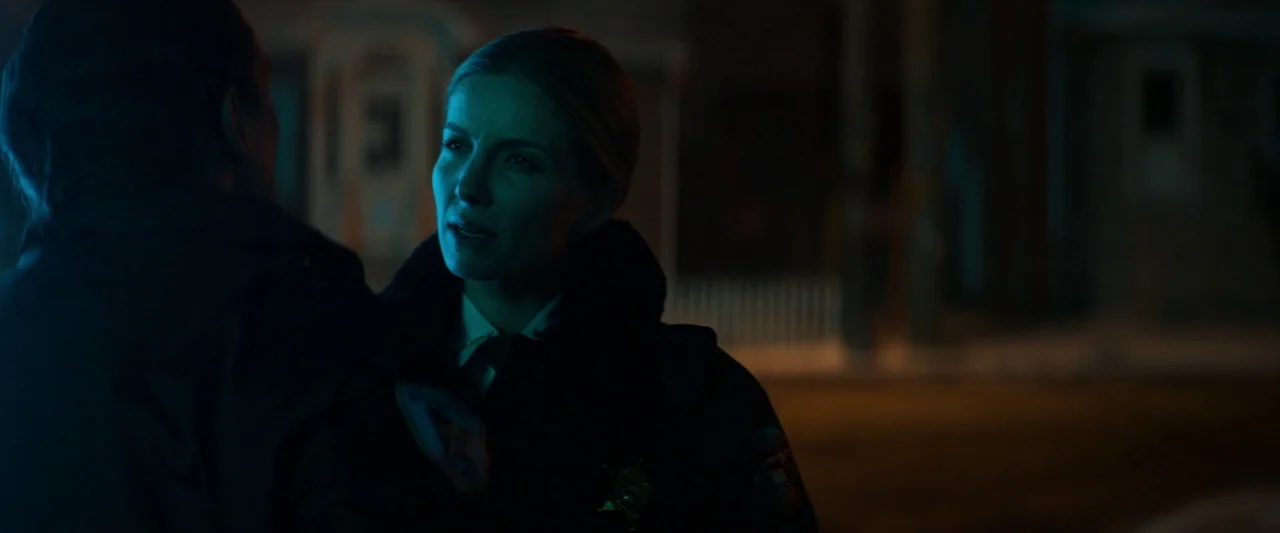 The Silencing (2020) movie still: medium — A dark residential street at night illuminated by distant yellow street lighting. A female police officer…