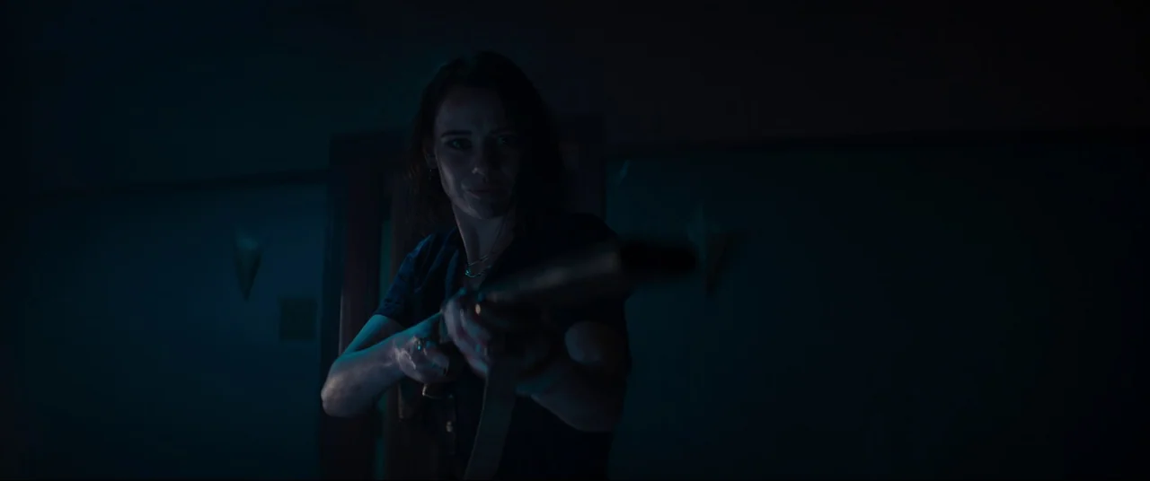Evil Dead Rise (2023) movie still: medium — A dark, dimly lit interior room features faint wall mounted sconces. A woman stands holding a shotgun,…