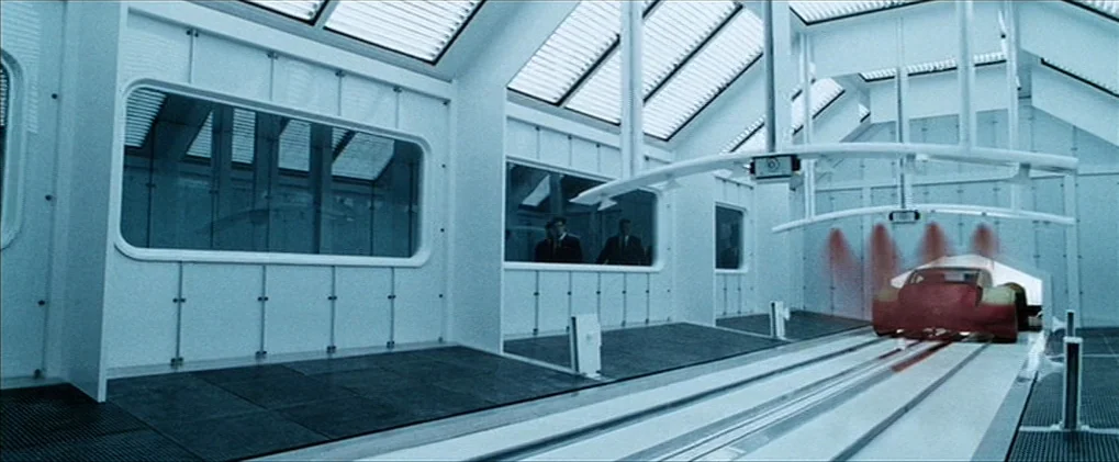 Minority Report movie still: wide — A stark white, clinical industrial chamber contains elevated observation windows along one side. A sleek red…