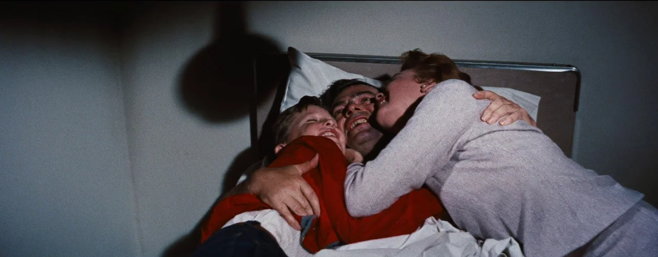 Bigger Than Life (1956) movie still: medium — A dark bedroom shows a family of three nestled together on a mattress. The parents and child are…
