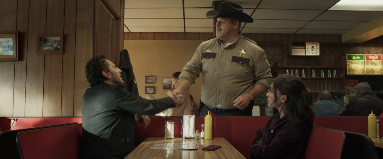 Clown in a Cornfield (2025) movie still: medium — A diner booth with wood-paneled walls, red vinyl seats, and a striped laminate table. A man in a…