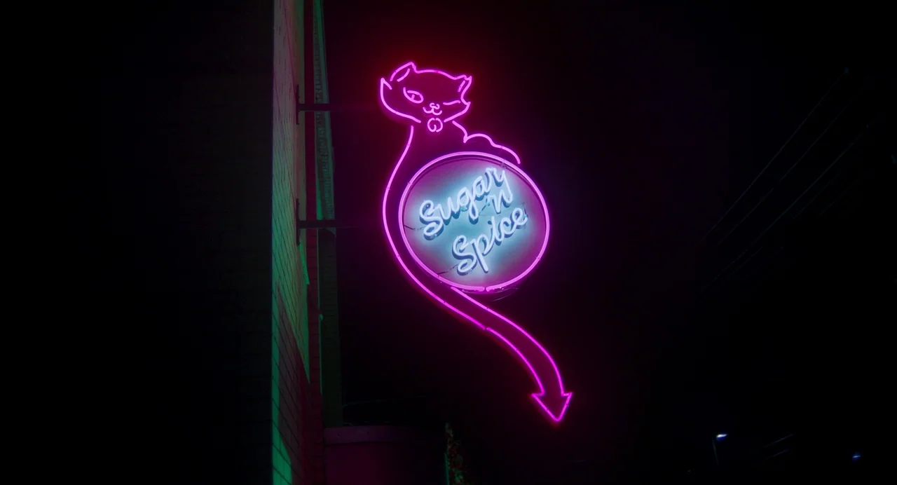 Drive-Away Dykes (2024) movie still: wide — A dark night exterior features a glowing neon sign mounted to a building wall. The sign depicts a…