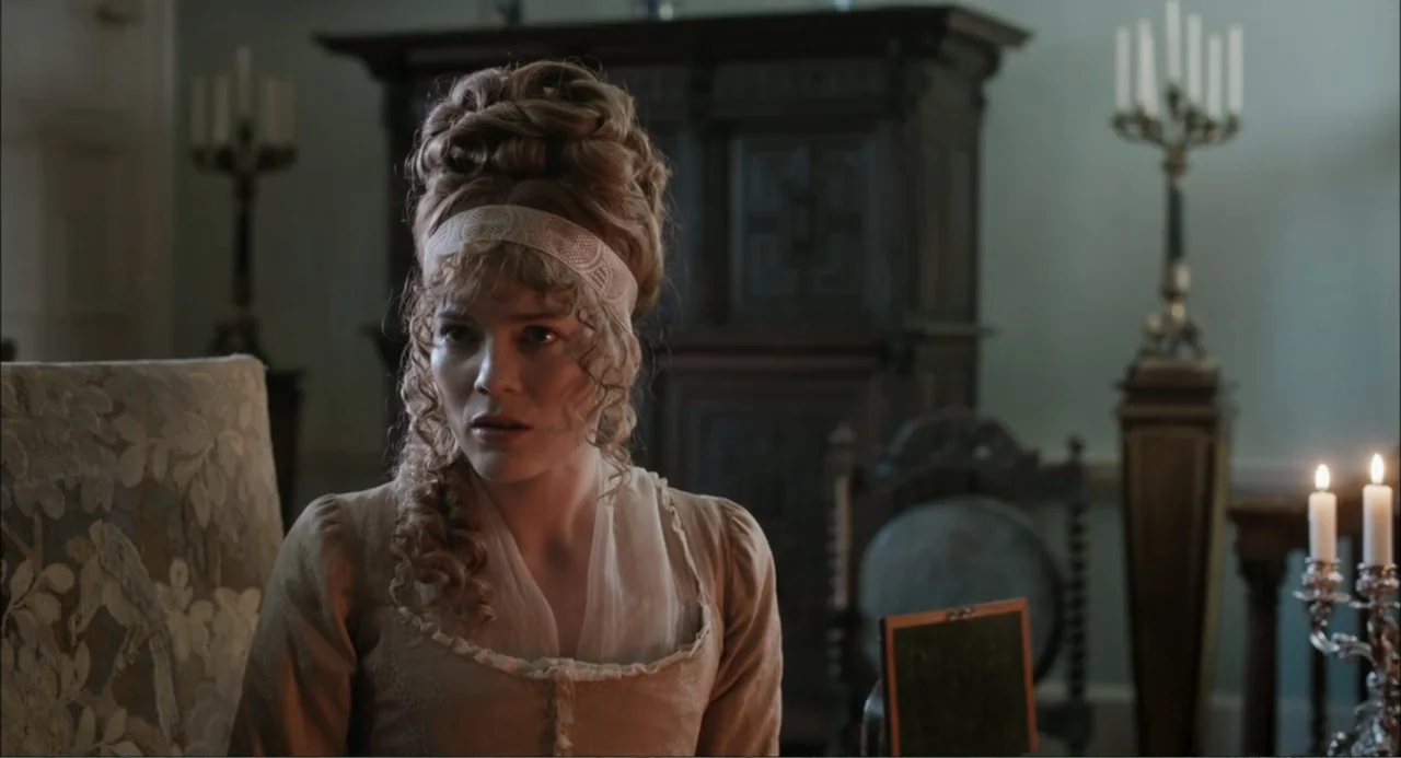Love & Friendship (2016) movie still: medium — A 19th-century parlor with an ornate wooden cabinet and candelabras in the background. A woman in…