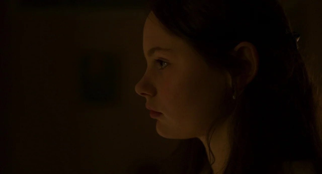 Zombi Child (2019) movie still: closeup — A dimly lit interior space with the background obscured by near-total darkness. A young girl's face is…