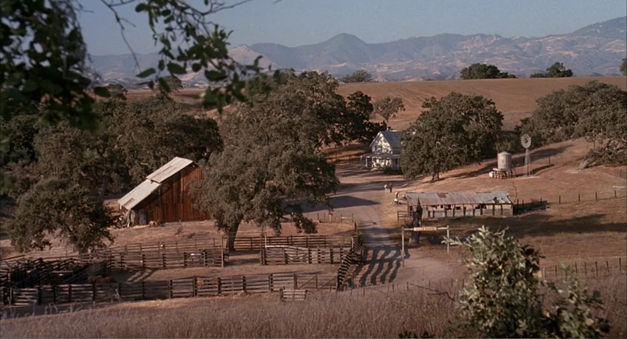 Of Mice and Men (1992) movie still: wide — A rural ranch property features a wooden barn, a white house, and livestock pens. The landscape is dotted…