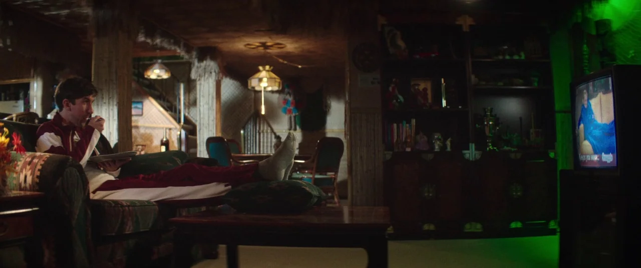 Room For Rent (2017) movie still: medium — A dimly lit room filled with mid-century furniture and wood paneling walls. A man rests on a patterned…