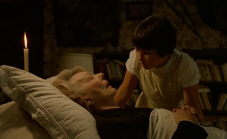 Milou en Mai (1990) movie still: medium — An elderly woman lies on a white pillow in a dimly lit, book-filled room. A young girl leans over the…