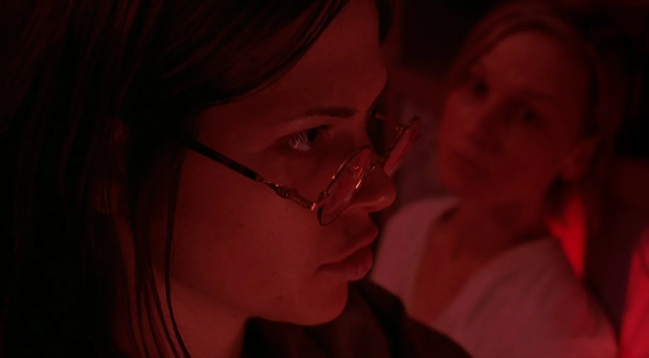 Cube (1997) movie still: closeup — Two women are positioned close together inside a dimly lit indoor space. The scene is bathed in pervasive red…