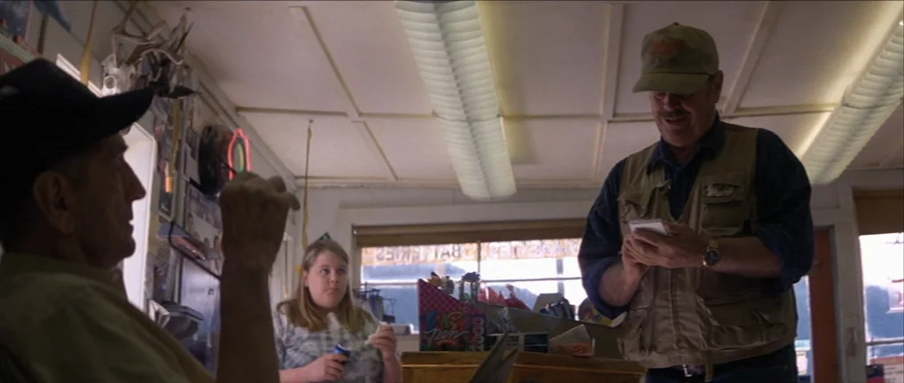 The Pledge (2001) movie still: medium — A small retail shop interior features a counter with displays and fluorescent ceiling panels. A man records…