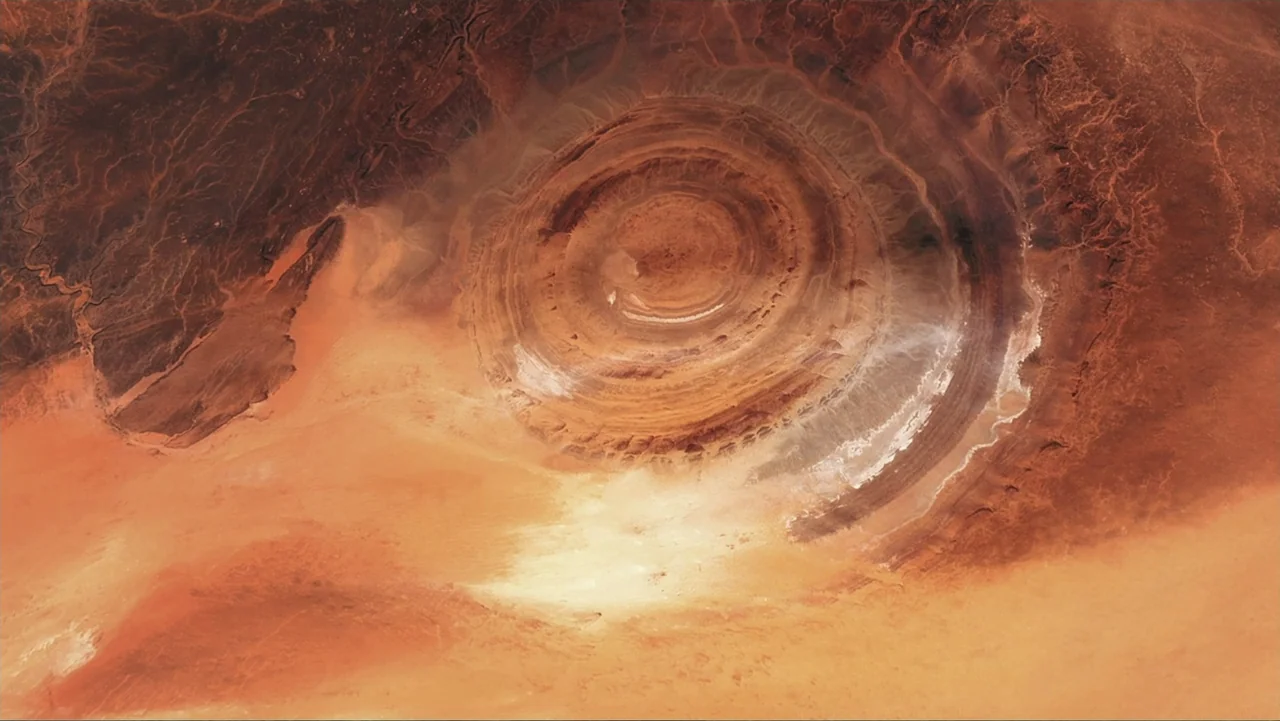 The Atlantis Puzzle (2024) movie still: establishing — A vast, arid desert landscape features a large, circular geological ring formation. No people…