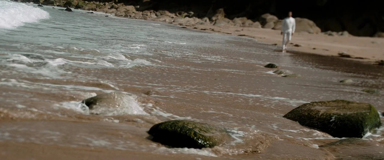Oxygen (2021) movie still: wide — A wet sand beach features scattered dark mossy rocks near the incoming surf. An adult male figure in all-white…