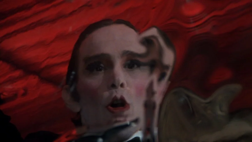 Cabaret (1972) movie still: closeup — A man looks into a rippled mirror inside a dark room with red walls. The reflection of his face is visibly…