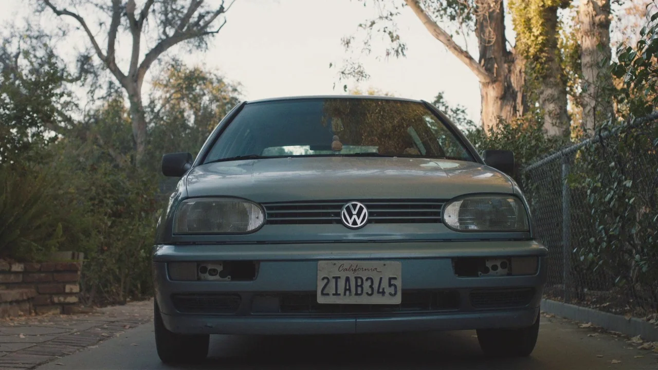 Beatriz at Dinner (2017) movie still: medium — A silver Volkswagen Golf sits parked on a concrete driveway near a chain-link fence. Mature trees…