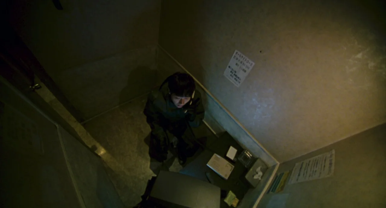 Ichi The Killer (2001) movie still: medium — A small, confined, sparse structure with plain walls and a table holding papers. A man sits alone…
